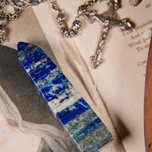 May include: A rectangular lapis lazuli crystal obelisk with blue and white marbling. A silver rosary and crucifix are in the background, along with a vintage print and a wooden object. The obelisk is a decorative item.