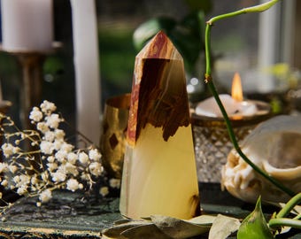 May include: A crystal obelisk with a yellow base and a brown and red top. The crystal is set against a backdrop of candles, flowers, and a skull, creating a mystical ambiance.