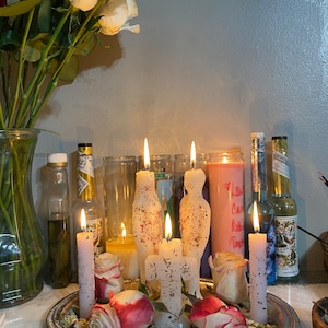 May include: A spiritual display featuring lit candles of various shapes and sizes, including figure-shaped candles, arranged on a silver tray with rose petals and dried herbs. A vase of red and white roses is on the left.