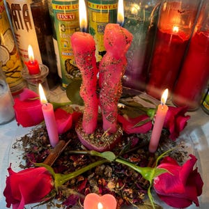 Könnte beinhalten: Zwei rosa figürliche Kerzen auf einem herzförmigen Sockel, umgeben von getrockneten Kräutern, vier rosa Rosen, zwei dünnen rosa Stabkerzen und einem kleinen herzförmigen Teelicht auf einem klaren Glasteller, mit hohen Glas-Stumpenkerzen.