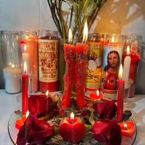 May include: A collection of lit candles, roses, and other decorative items. Several red candles are burning, along with candles in glass jars. A bouquet of roses sits in the background. The arrangement is on a clear glass plate.