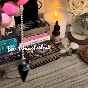 Puede incluir: Un péndulo negro con una cadena plateada cuelga sobre una pila de cartas de tarot. Las cartas están etiquetadas como "The Hidden Truth Oracle" y "Bewitching Parlour". Hay una vela encendida, una botella de aceite esencial y cristales sobre una mesa de madera.