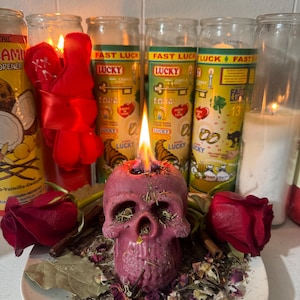 May include: A burning, pink skull candle rests on a white plate with dried herbs, rose petals, and two red roses. Several glass jar candles with the text "FAST LUCK" are in the background, along with a red ribbon.