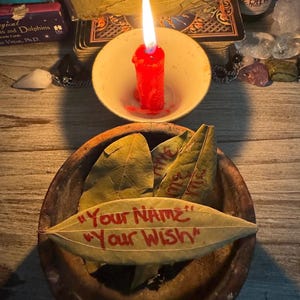 May include: A lit red candle in a white bowl sits above a wooden bowl with green leaves. One leaf has the text "Your NAME" and "Your Wish" in red. Tarot cards and a bottle are in the background.