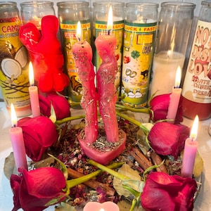 May include: A spiritual arrangement featuring lit candles, including a heart-shaped candle and tall pink ones. Two intertwined red candles are central, surrounded by red roses, herbs, and cinnamon sticks on a glass plate. Various glass jar candles are in the background.