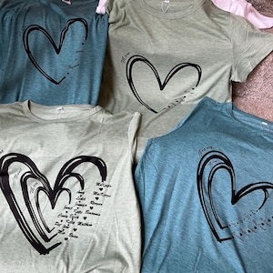 May include: A selection of t-shirts in shades of teal, green, and white. Each shirt has a large black heart design, with names and titles such as "Mom" and "Nana" printed inside. The shirts are made from a soft fabric.