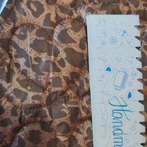 May include: Close-up of a brown and tan leopard print fabric with a quilted pattern. A white ruler with blue illustrations and the word "Handmade" is on the right side of the image. The fabric is likely used for sewing or crafting projects.