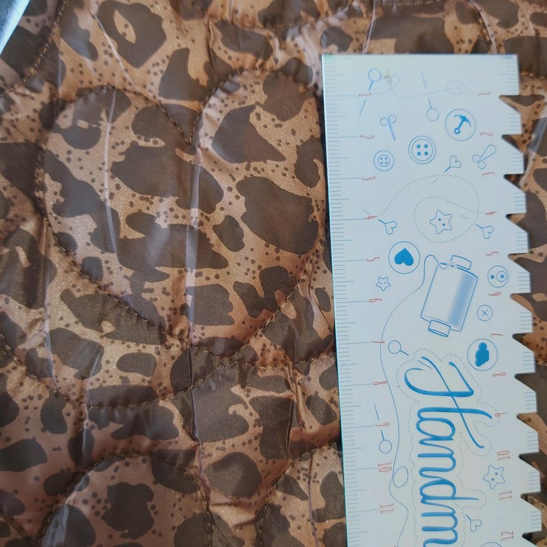 May include: Close-up of a brown and tan leopard print fabric with a quilted pattern. A white ruler with blue illustrations and the word "Handmade" is on the right side of the image. The fabric is likely used for sewing or crafting projects.