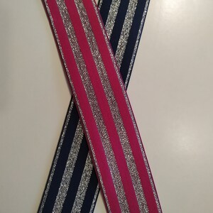 K&ouml;nnte beinhalten: Zwei elastische B&auml;nder, eines in Pink und eines in Marineblau, kreuzen sich in X-Form. Beide B&auml;nder haben vertikale silberfarbene Metallic-Streifen. Die B&auml;nder werden wahrscheinlich zum N&auml;hen, Basteln oder zur Herstellung von Kleidungsst&uuml;cken verwendet.