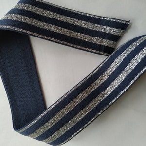 K&ouml;nnte beinhalten: Marineblaues elastisches Band mit silbernen Glitzerstreifen. Das Band ist &uuml;ber sich selbst gefaltet und zeigt beide Seiten des Materials. Die Streifen sind gleichm&auml;&szlig;ig verteilt und verlaufen parallel zueinander. Das Band wird wahrscheinlich f&uuml;r N&auml;h- oder Bastelprojekte verwendet.