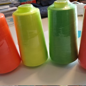 Könnte beinhalten: Vier Garnrollen in leuchtenden Farben: Orange, Gelb, Grün und Pink. Die Rollen sind in durchsichtige Plastikfolie verpackt.