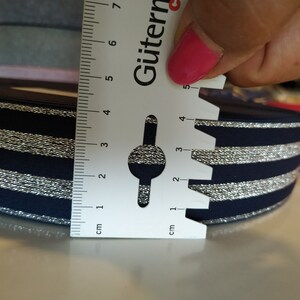 K&ouml;nnte beinhalten: Marineblaues Band mit silbernen Metallic-Streifen. Das Band ist etwa 4 cm breit. Ein Lineal wird auf das Band gelegt, um seine Breite zu messen. Das Band liegt auf einer wei&szlig;en Oberfl&auml;che.