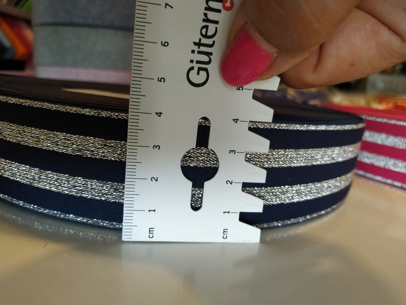 K&ouml;nnte beinhalten: Marineblaues Band mit silbernen Metallic-Streifen. Das Band ist etwa 4 cm breit. Ein Lineal wird auf das Band gelegt, um seine Breite zu messen. Das Band liegt auf einer wei&szlig;en Oberfl&auml;che.
