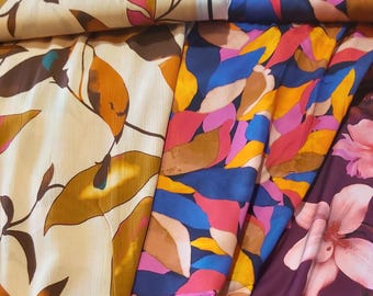 May include: Three fabric swatches with different patterns. The first swatch is a cream fabric with a brown leaf pattern. The second swatch is a blue fabric with a colorful abstract leaf pattern. The third swatch is a purple fabric with a pink flower pattern.