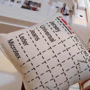 Könnte beinhalten: Ein weißer Nähmaschinenfuß mit einem Raster aus Zahlen und Linien, das darauf gedruckt ist. Der Fuß ist mit verschiedenen Stoffarten beschriftet, darunter "Jersey", "Universal", "Jeans", "Leder" und "Microtex".
