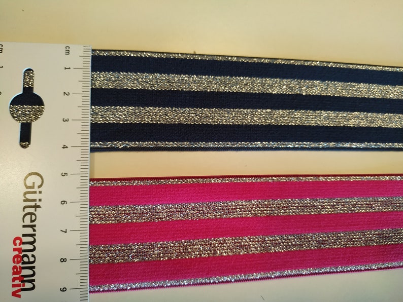 K&ouml;nnte beinhalten: Zwei Streifen gestreiftes elastisches Band. Einer ist marineblau mit silbernen Streifen, der andere pink mit silbernen Streifen. Ein Lineal liegt neben den B&auml;ndern, mit Messungen in Zentimetern.