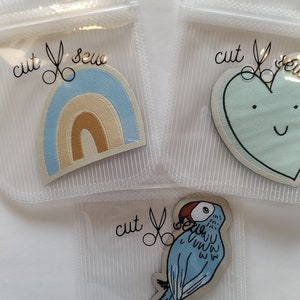 Könnte beinhalten: Drei Bügelbilder in durchsichtigen Plastiktüten. Die Bilder sind ein Regenbogen, ein Herz mit einem Smiley und ein blauer Vogel. Jede Tüte trägt den Text "cut & sew" mit einer Schere.