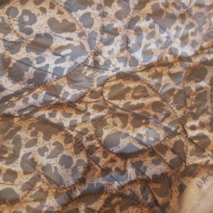 May include: Close-up of a quilted fabric with a leopard print pattern. The fabric features a tan background with dark gray spots, stitched in a diamond pattern. The overall texture is smooth and slightly shiny.