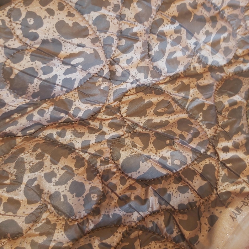 May include: Close-up of a quilted fabric with a leopard print pattern. The fabric features a tan background with dark gray spots, stitched in a diamond pattern. The overall texture is smooth and slightly shiny.