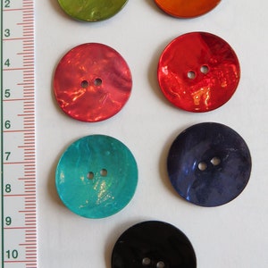 Könnte beinhalten: Sechs runde, zweilöcherige Knöpfe in verschiedenen Farben, darunter Grün, Orange, Rot, Blau, Türkis und Schwarz. Die Knöpfe sind aus einem glänzenden, irisierenden Material gefertigt.