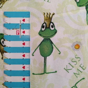 Könnte beinhalten: Ein weißer Stoff mit einem grünen Frosch, der eine goldene Krone trägt und den Text "Kiss Me". Der Stoff hat ein sich wiederholendes Muster aus Fröschen, Blumen und dem Text "Kiss Me".