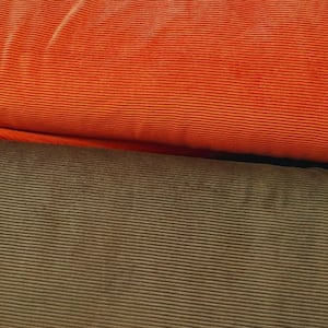 Könnte beinhalten: Nahaufnahme von zwei Cordstoffen. Der obere Stoff ist ein leuchtendes Orange, während der untere Stoff ein gedämpftes Olivgrün ist. Beide Stoffe haben eine gerippte Textur, wobei die Rippen horizontal verlaufen. Der orangefarbene Stoff ist über dem grünen Stoff gefaltet.