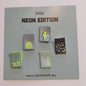 Könnte beinhalten: Ein Set mit fünf Bügelbildern mit neongelben Designs auf grauem Hintergrund. Die Designs beinhalten einen Blitz, ein Smiley, das Wort "YAY!" und das Wort "YEAH!" Die Patches werden auf einem hellgrünen Hintergrund mit dem Text "LALELOUP NEON EDITION" und "www.LALELOUP.de" angezeigt.
