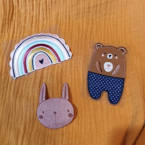 Könnte beinhalten: Drei Filzpatches, ein Regenbogen mit einem Herzen, ein brauner Bär mit blauer Polka-Dot-Hose und ein braunes Kaninchen.