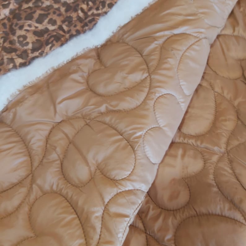 May include: A tan quilted blanket with a heart pattern. The blanket has a soft, plush white border and a leopard print fabric on the reverse side. The blanket is folded, showing the heart pattern and the leopard print.