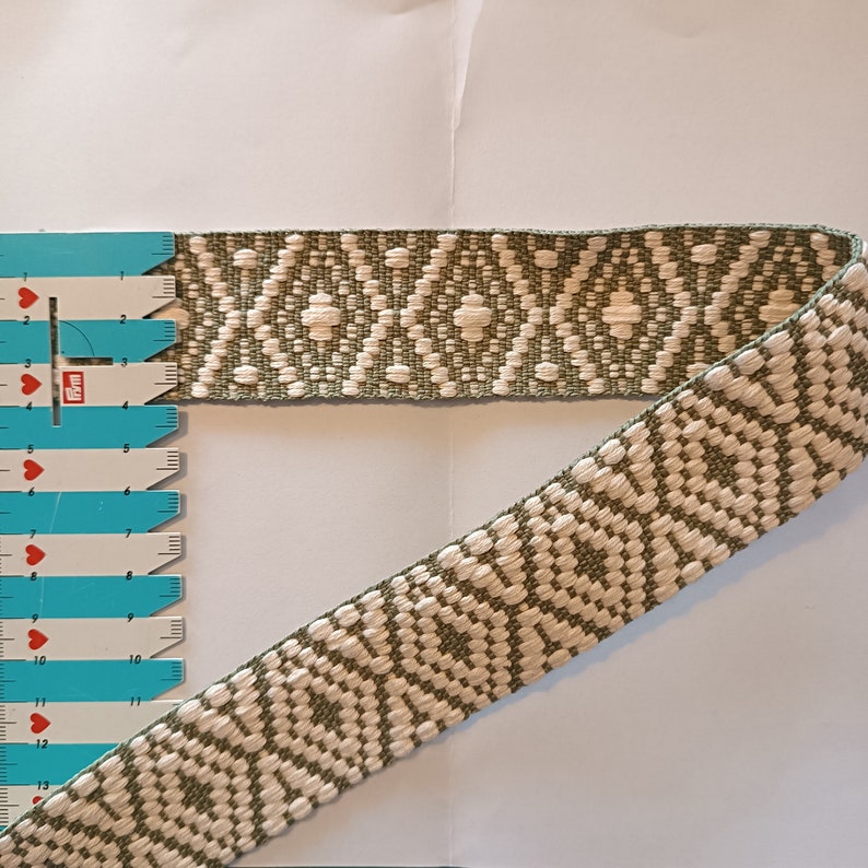 Puede incluir: Cinta tejida con un patr&oacute;n geom&eacute;trico de diamantes en blanco roto y verde oliva. Se utiliza una regla azul y blanca para medir la cinta. La cinta es adecuada para coser, manualidades o fines decorativos.