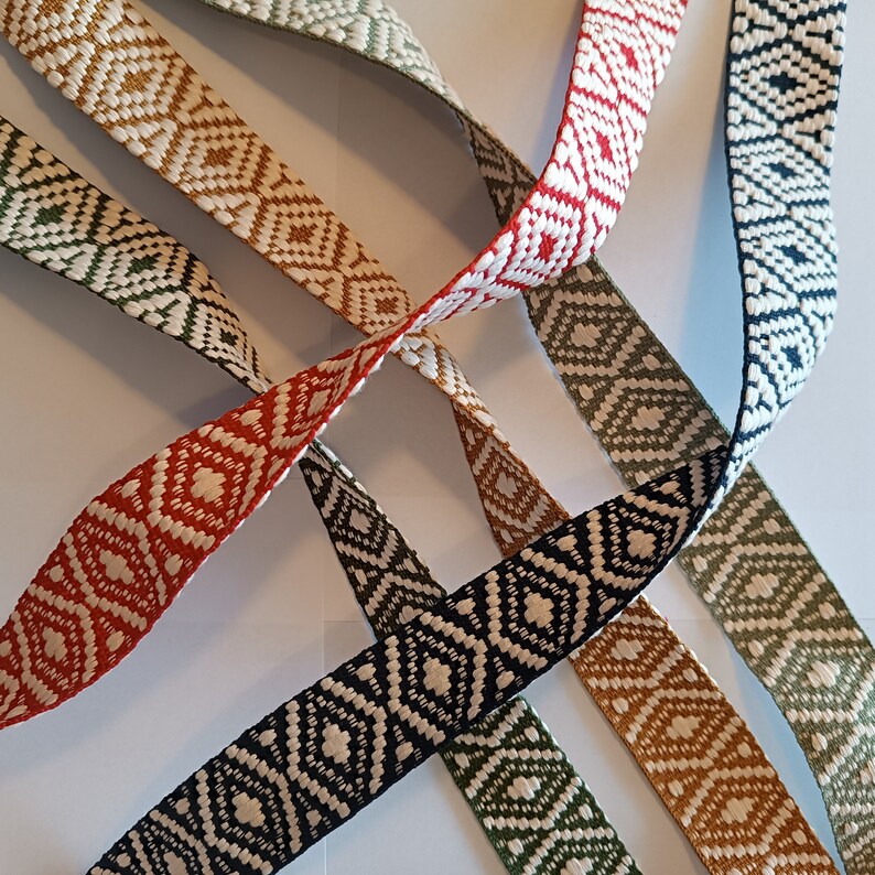 Puede incluir: Primer plano de varias cintas tejidas en varios colores, incluyendo rojo, blanco, verde y marr&oacute;n. Cada cinta presenta un patr&oacute;n de diamantes repetido. Las cintas est&aacute;n dispuestas en un patr&oacute;n entrecruzado sobre un fondo blanco.