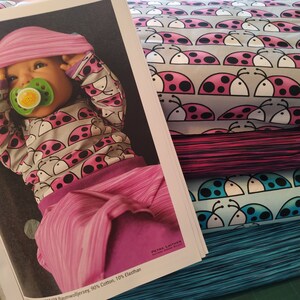 Könnte beinhalten: Stapel von Stoffrollen mit einem pink-schwarzen Marienkäfermuster auf grauem Hintergrund und ein Schnittmusterbuch. Das Buch zeigt ein Baby in einem passenden Outfit. Ideal für Kinderkleidung.