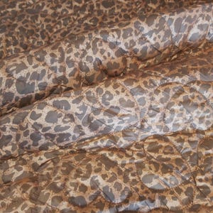 May include: Close-up of a quilted fabric with a leopard print pattern. The fabric features a repeating design of brown and tan spots on a lighter tan background. The quilting creates a diamond pattern.