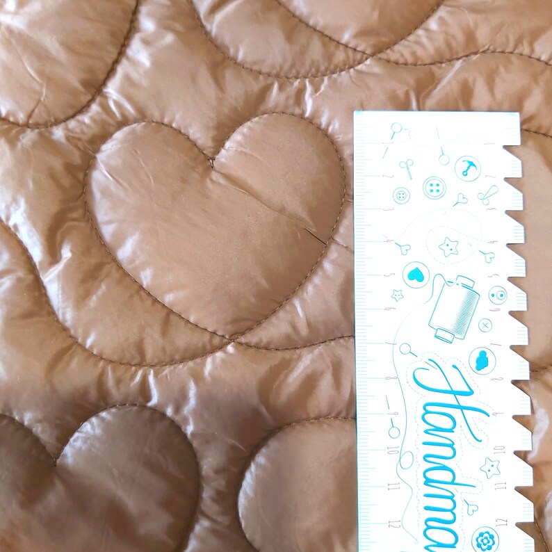 May include: Close-up of a quilted fabric with heart-shaped patterns in a light brown color. A white ruler with blue illustrations and the word "Handmade" is on the right side of the image.