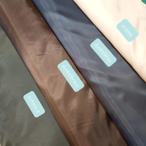 Könnte beinhalten: Nahaufnahme von Stoffmustern in verschiedenen Farben. Die Farben umfassen Dunkelgrau, Braun, Marineblau und Creme. Jedes Stoffmuster hat ein Etikett mit einem deutschen Farbnamen. Die Stoffe scheinen glatt und glänzend zu sein.