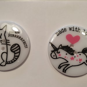Könnte beinhalten: Zwei weiße, runde Knöpfe mit schwarzen und rosa Grafiken. Ein Knopf zeigt eine Katze mit dem Text "you are purrrfect!". Der andere Knopf hat ein Einhorn mit einem rosa Herz und den Worten "made with love".