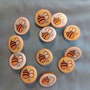 Könnte beinhalten: Eine Sammlung von 11 runden, bunten Knöpfen mit Bienenmotiv. Die Knöpfe sind mit gelben und schwarzen Bienen verziert, einige haben einen weißen Hintergrund mit einem gelben und schwarzen Bienenmotiv. Die Knöpfe sind perfekt, um jedem Projekt eine Prise Verspieltheit zu verleihen.