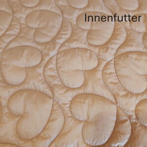 May include: Close-up of a quilted fabric with a heart-shaped pattern in a light beige color. The fabric has a satin sheen and the text "Innenfutter" is visible.
