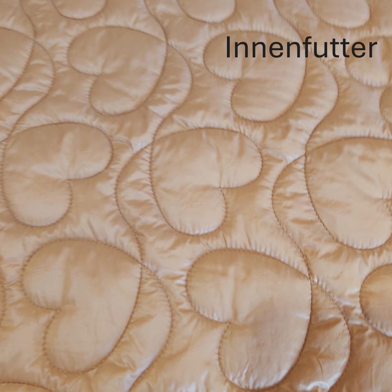 May include: Close-up of a quilted fabric with a heart-shaped pattern in a light beige color. The fabric has a satin sheen and the text "Innenfutter" is visible.