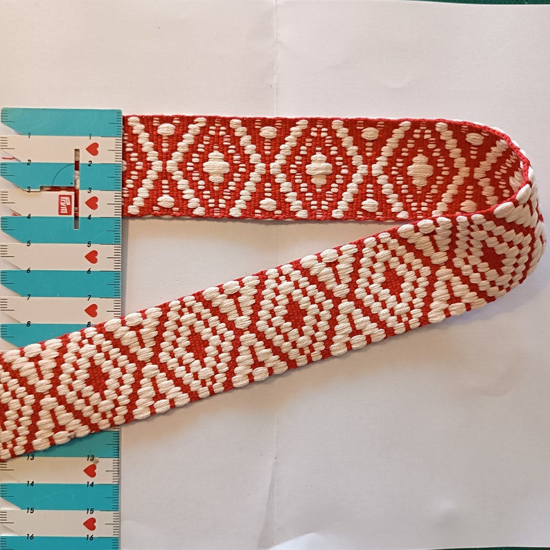 Puede incluir: Cinta tejida con un patr&oacute;n de diamantes rojos y blancos. La cinta, medida con una regla, tiene aproximadamente 3,8 cm de ancho. La cinta se utiliza probablemente para manualidades o fines decorativos.