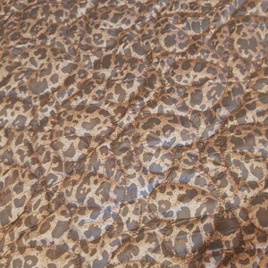 May include: A quilted fabric with a leopard print pattern. The fabric features a repeating design of brown spots on a tan background. The quilting creates a textured, padded appearance. The overall color palette is neutral, with shades of brown and tan.