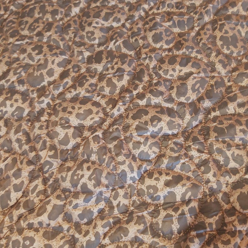 May include: A quilted fabric with a leopard print pattern. The fabric features a repeating design of brown spots on a tan background. The quilting creates a textured, padded appearance. The overall color palette is neutral, with shades of brown and tan.