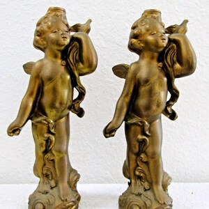 May include: A pair of gold cherub candle holders. Each cherub is holding a bird in its hand and is standing on a cloud-like base.