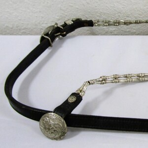 May include: A black leather belt with silver chain accents and two large silver conchos. The belt is laid out flat on a white surface.