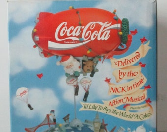 May include: A Coca-Cola themed musical Christmas decoration featuring a red blimp with the Coca-Cola logo, Santa Claus, a reindeer, and a globe with landmarks. The text "Delivered by the NICK in time Action Musical" is on the box.
