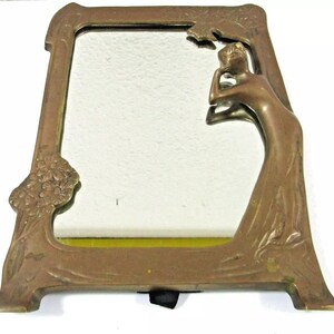 May include: A brass mirror with an ornate frame featuring a stylized female figure. The frame is decorated with floral motifs and the mirror has a beveled edge.