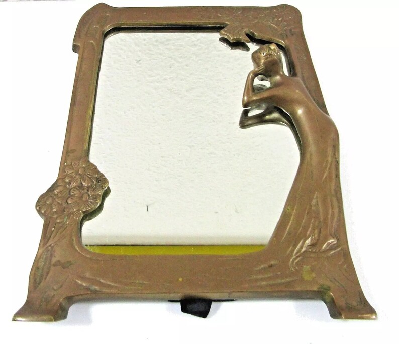 May include: A brass mirror with an ornate frame featuring a stylized female figure. The frame is decorated with floral motifs and the mirror has a beveled edge.