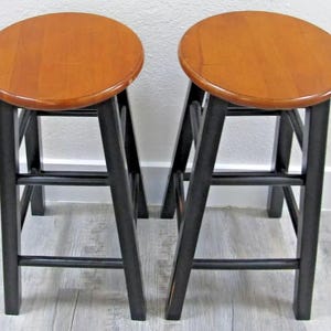 May include: Two wooden bar stools with black painted legs and a natural wood seat. The stools have a simple design with a single rung on each side.