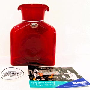 May include: A vibrant red Blenko glass vase with a square base and a flared top. The vase has a silver-colored metal band around the neck and a Blenko Handcraft sticker. A card with the text "Enjoy Your Piece of History in the Making!" is also present.