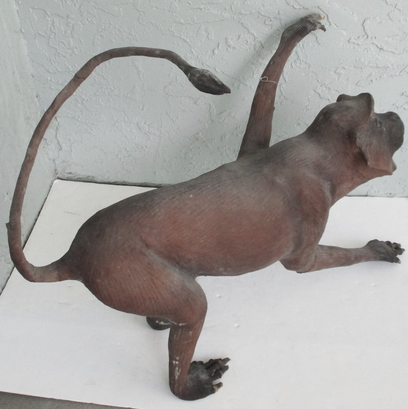 May include: A brown, taxidermied monkey figurine with a long tail curled behind it. The monkey is posed in a sitting position with its right arm raised and its left arm extended forward.